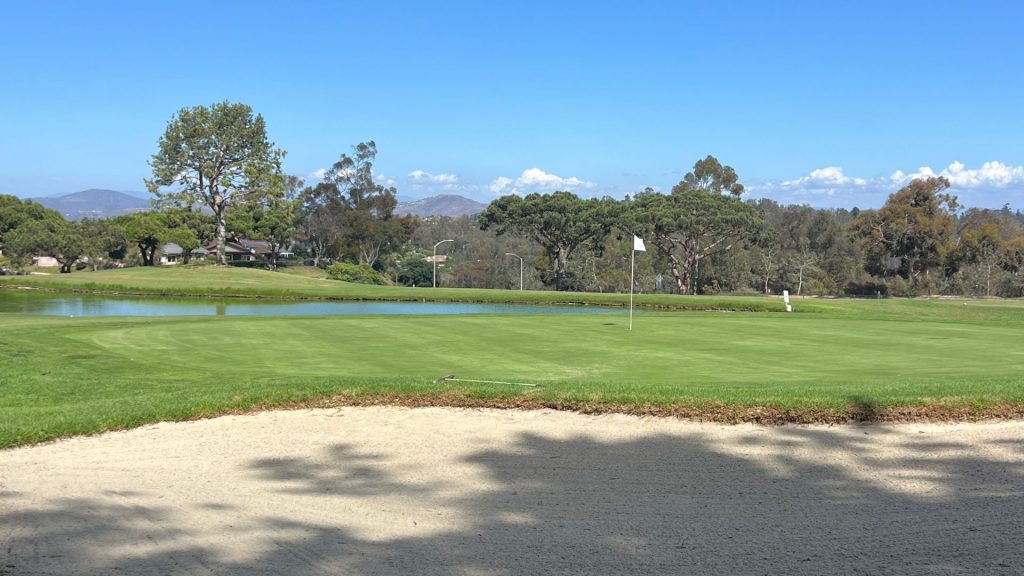 Putting green beside a calm pond with a greenside bunker and distant mountains at Lomas Santa Fe golf course