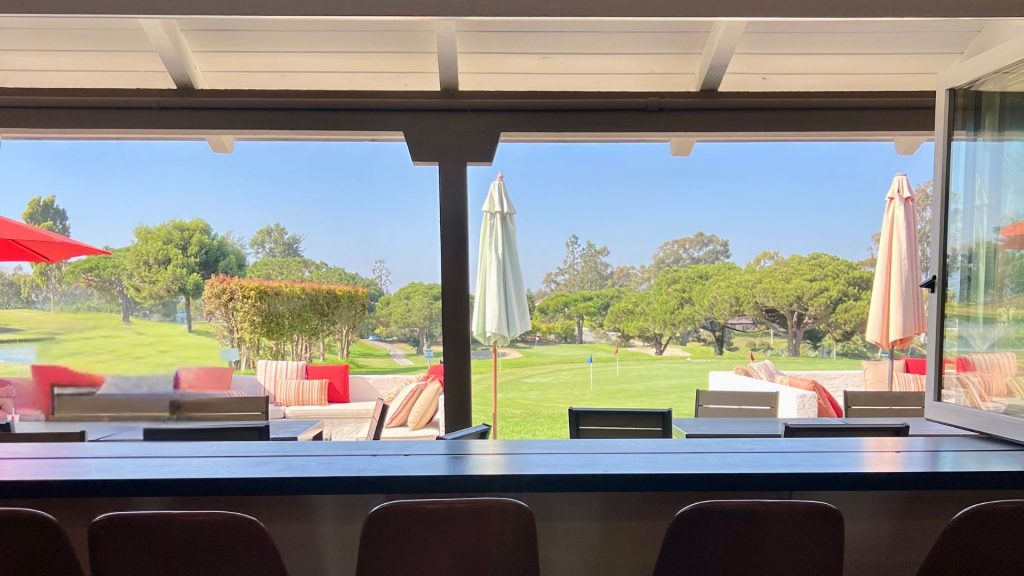 View from the Lomas Santa Fe Executive clubhouse bar across an outdoor patio with lounge seating to the practice putting green